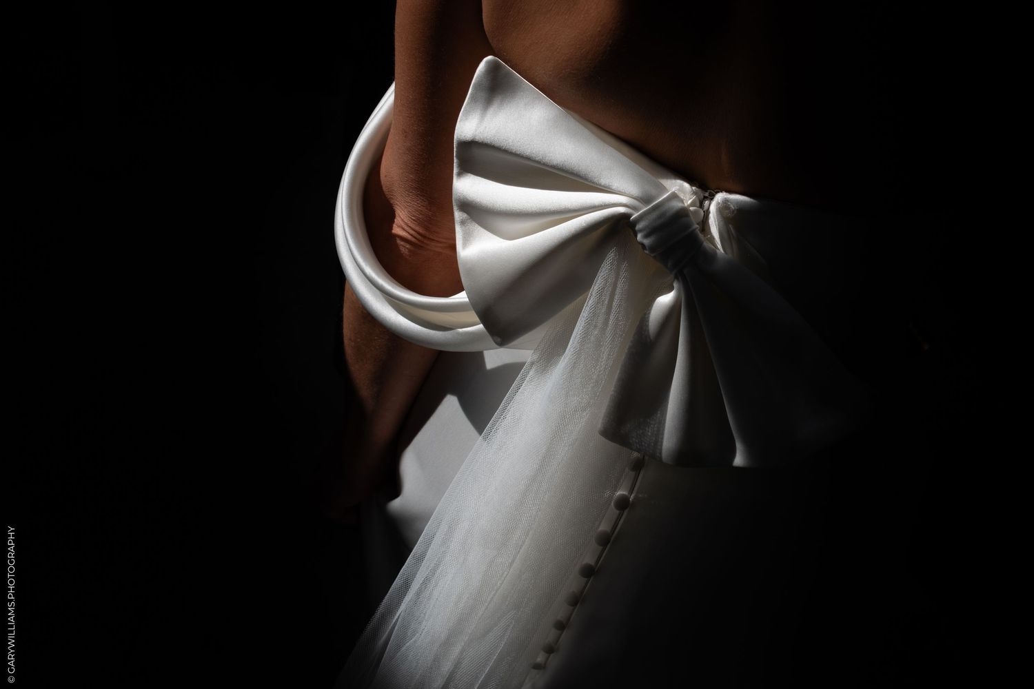 Close-up of a bride's white wedding dress bow detail against a dark background.