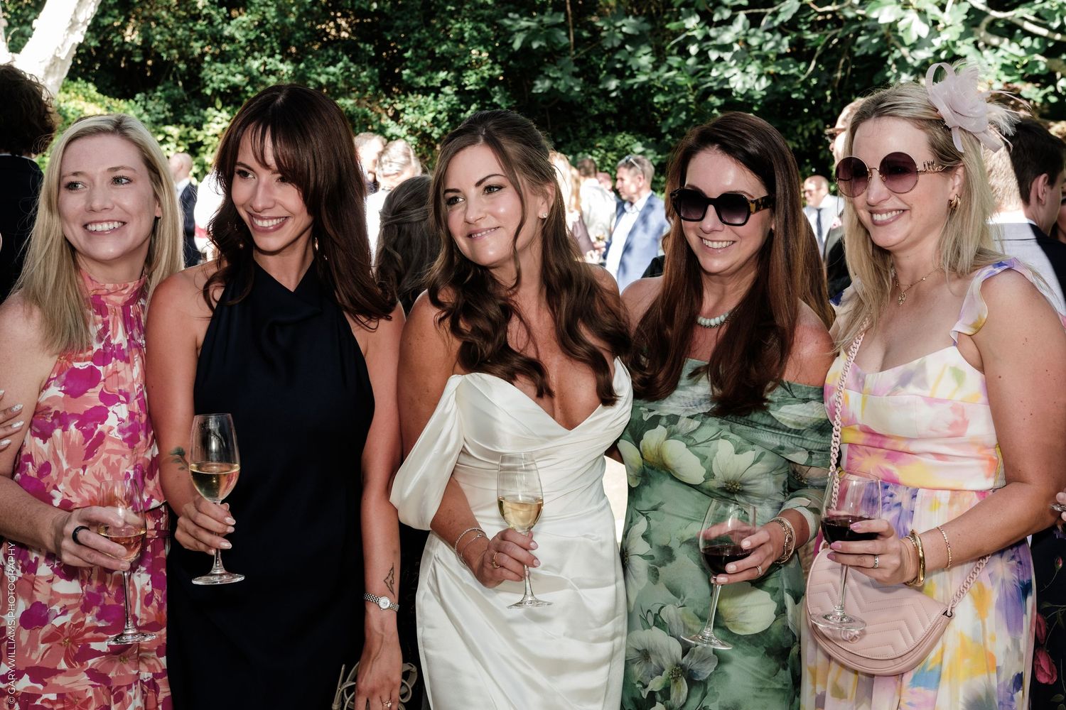 Five women smiling and holding wine glasses at an outdoor garden party event.