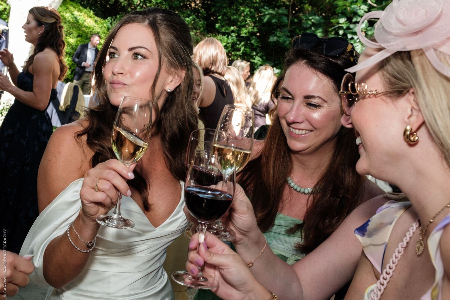 Three women in elegant attire laughing and toasting with wine glasses at an outdoor wedding reception.