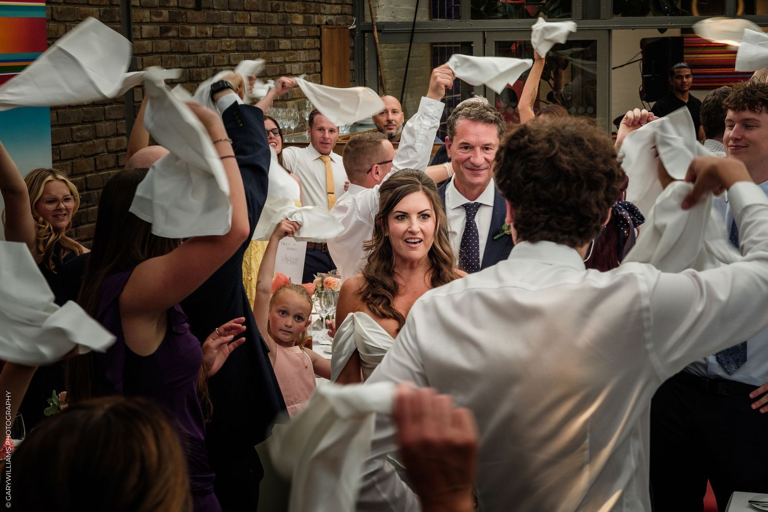 Bride and groom celebrate with guests waving white napkins at a lively indoor wedding reception party.