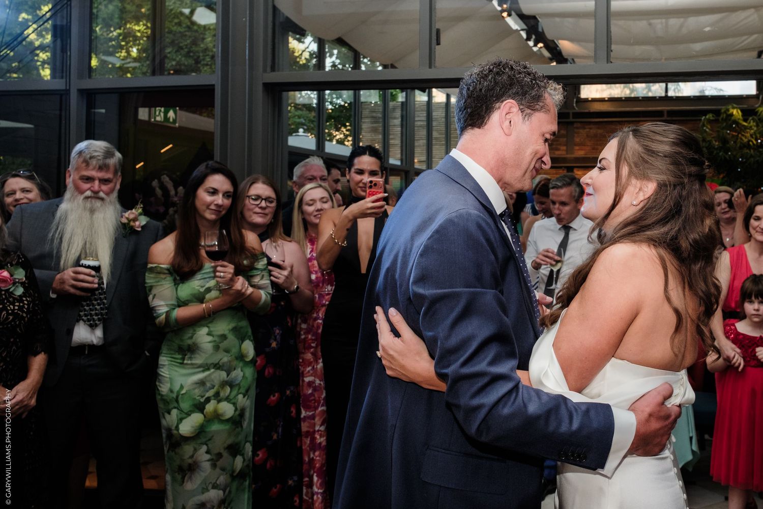 Bride and groom share first dance surrounded by smiling wedding guests at elegant indoor reception venue.