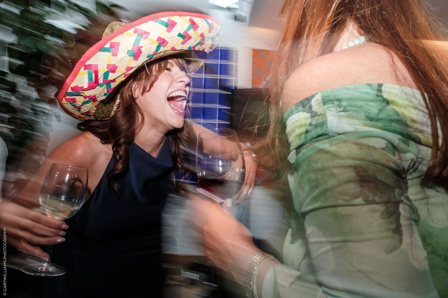 Woman in colorful hat laughing with wine glass at lively party, dancing near woman in green floral dress.
