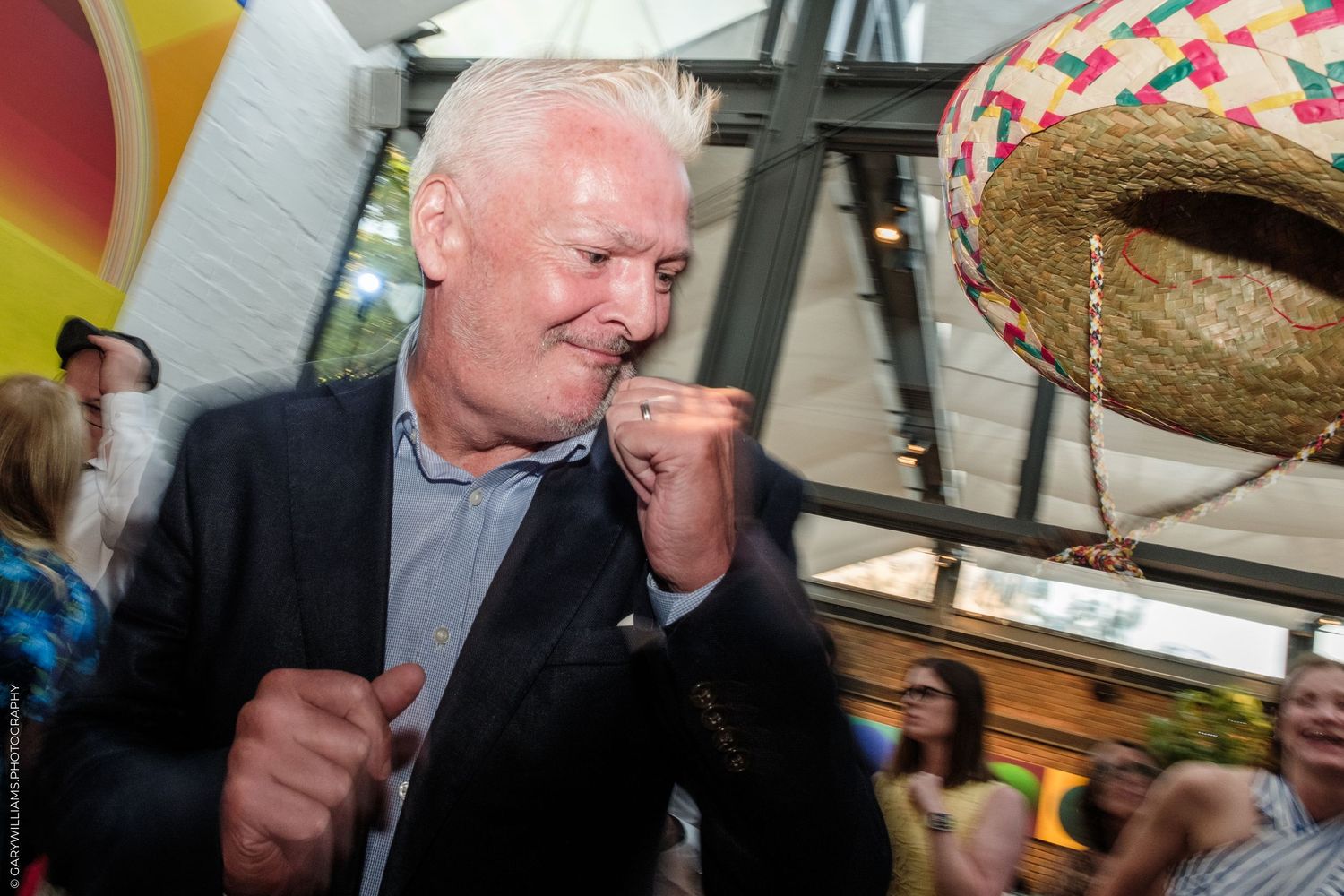 Man in blazer celebrating with fists raised at colorful outdoor party with sombrero decoration.