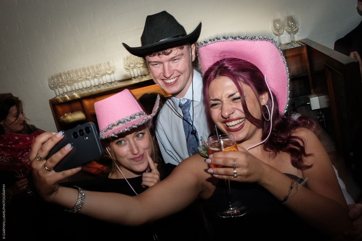 Three people posing for a selfie at a party, two women wearing pink cowboy hats and a man in a black cowboy hat.
