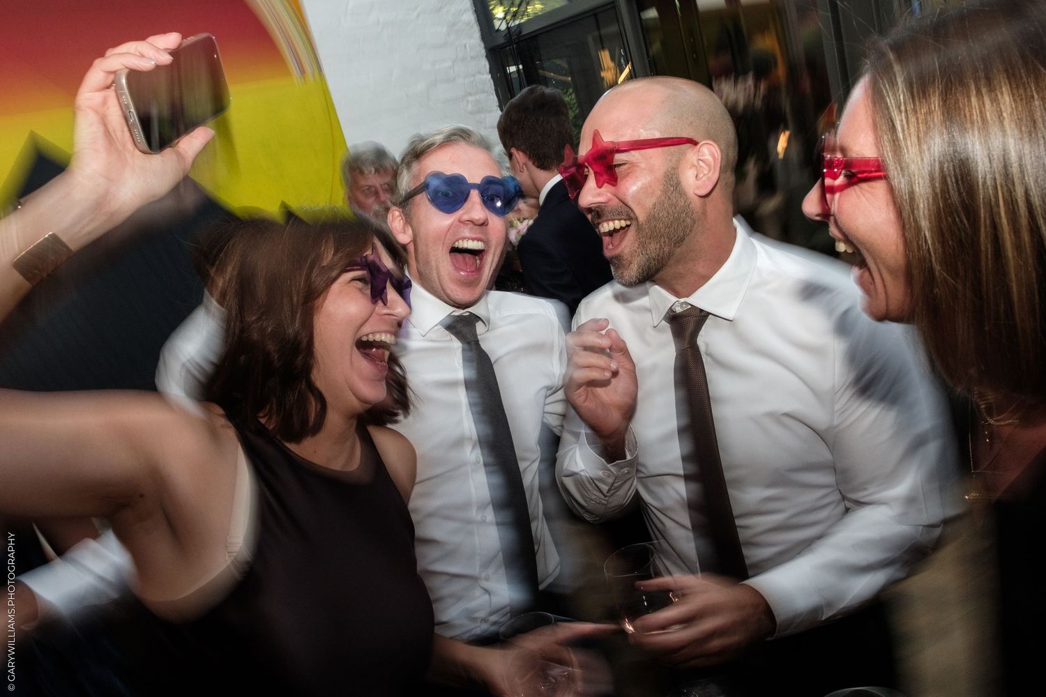 Group of friends laughing and dancing at a party, wearing colorful sunglasses and celebrating together.