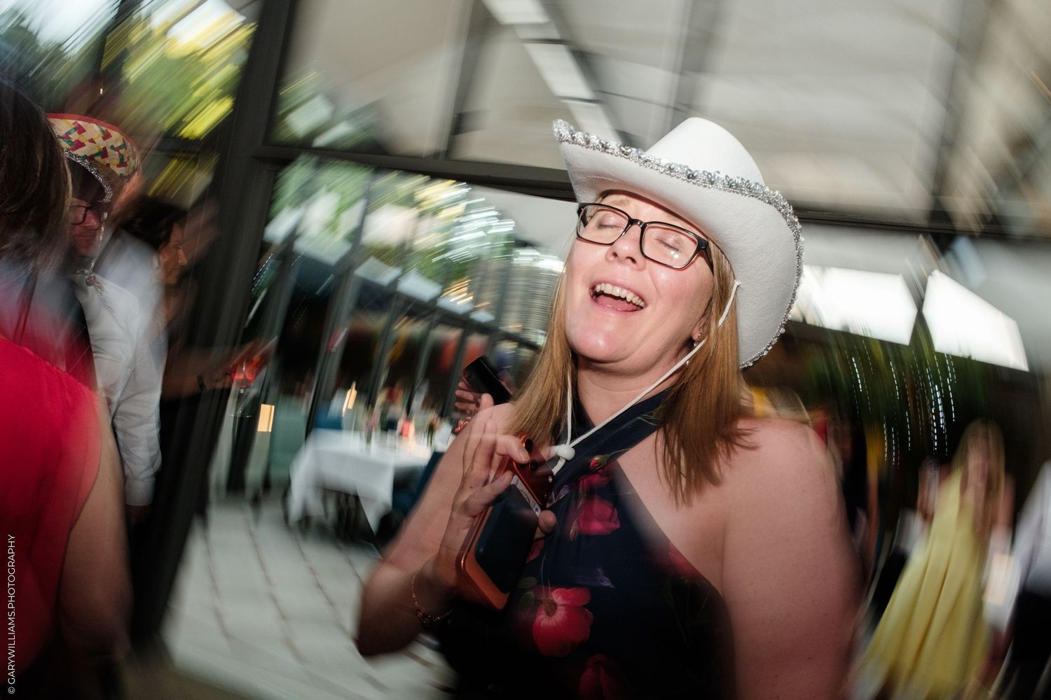 Woman in white cowboy hat and glasses laughing joyfully at an outdoor party event.