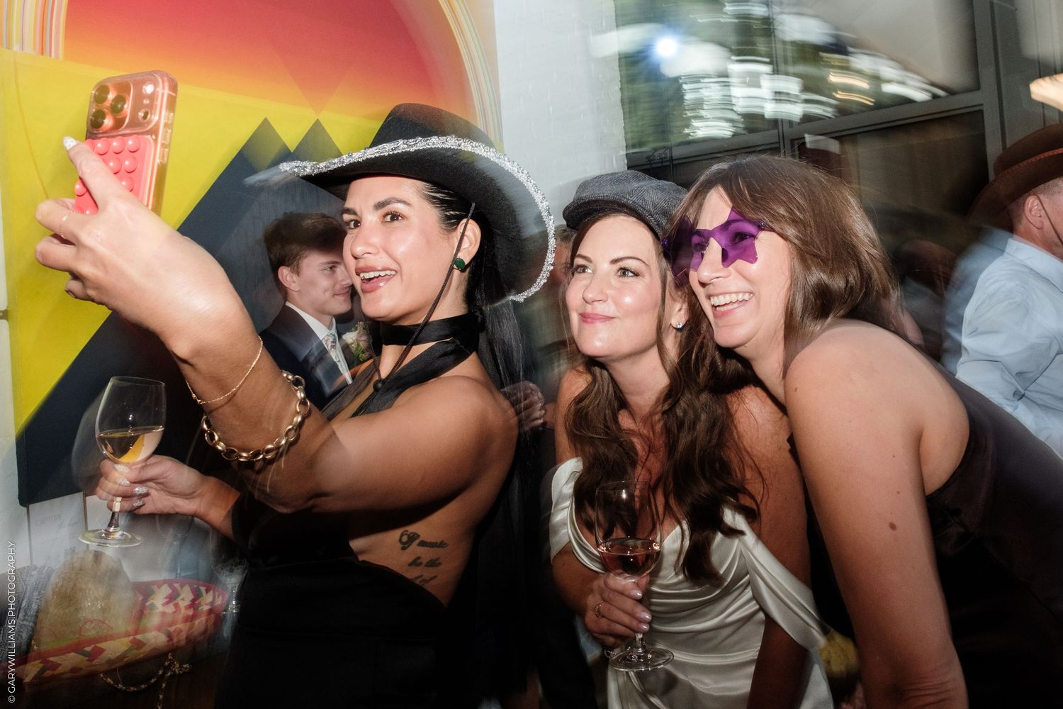 Three women taking a selfie at a party, wearing hats and holding drinks, smiling and having fun.