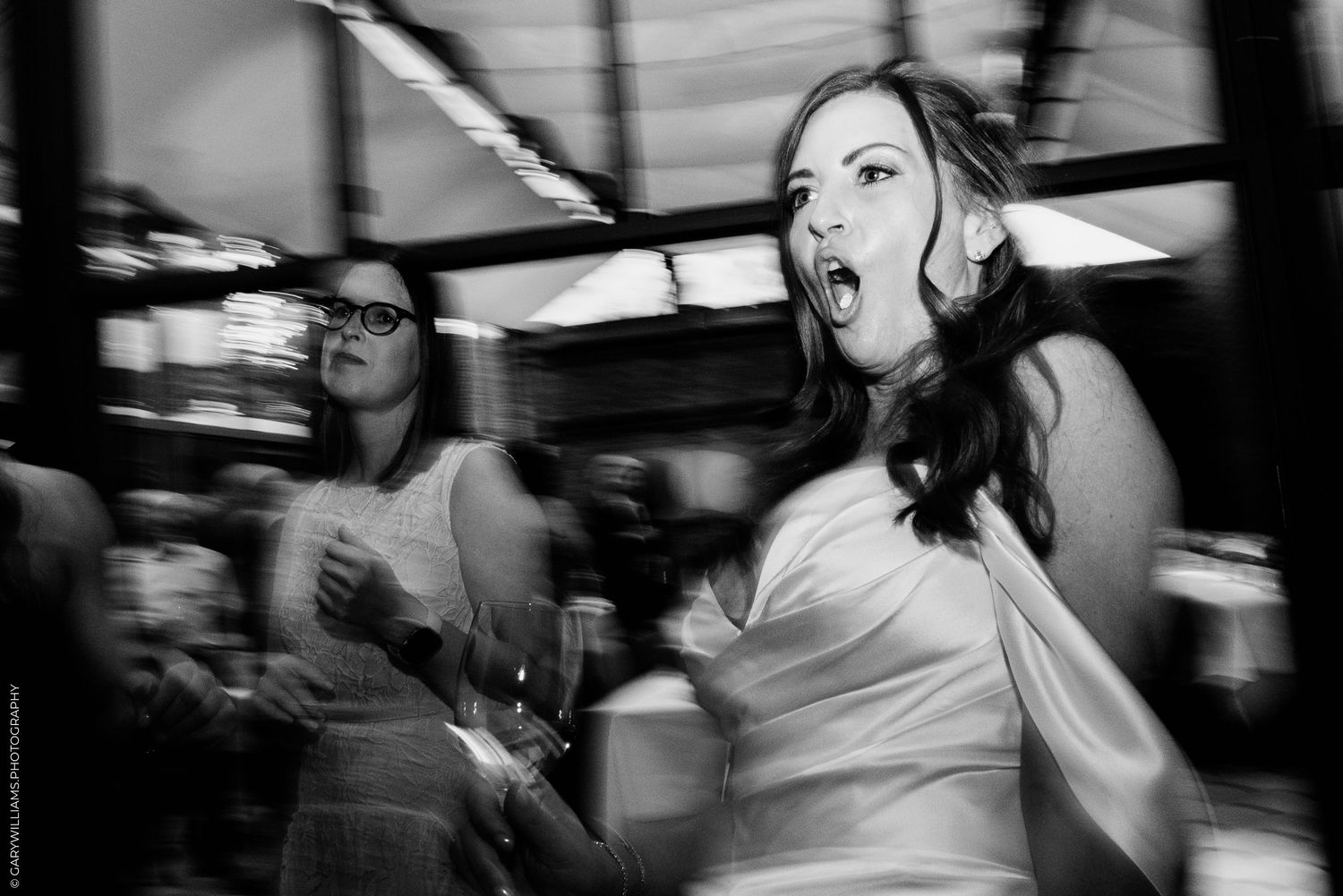 Black and white photo of a woman dancing expressively at a wedding reception with guests in the background.