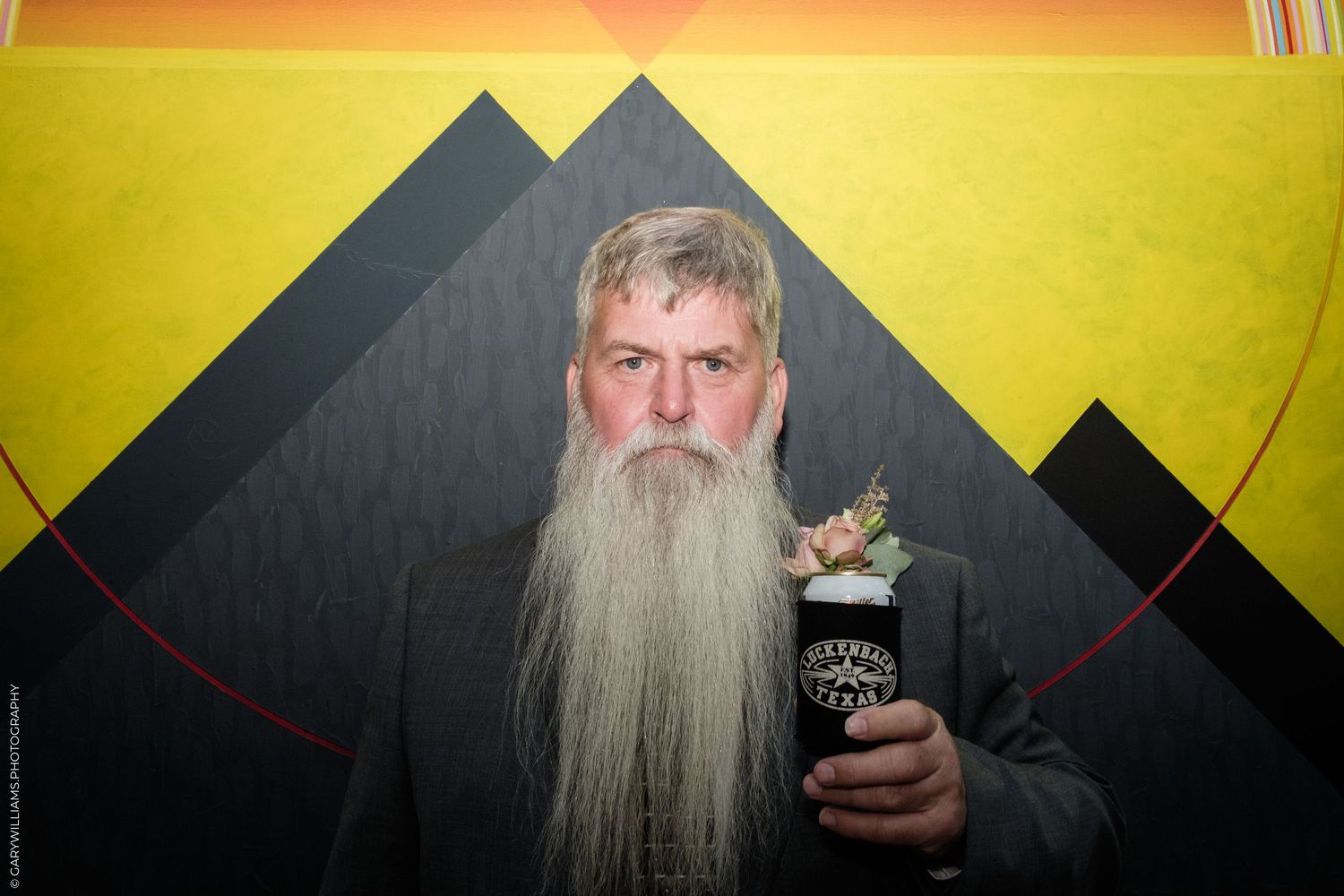 Elderly man with long gray beard holding a decorated cup against a geometric yellow and black background.