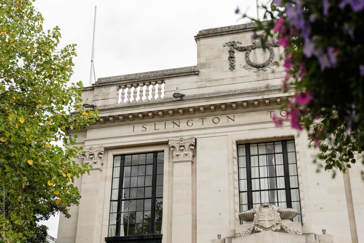 Islington Town Hall neoclassical stone facade with ornate details, framed by green trees and purple flowers.