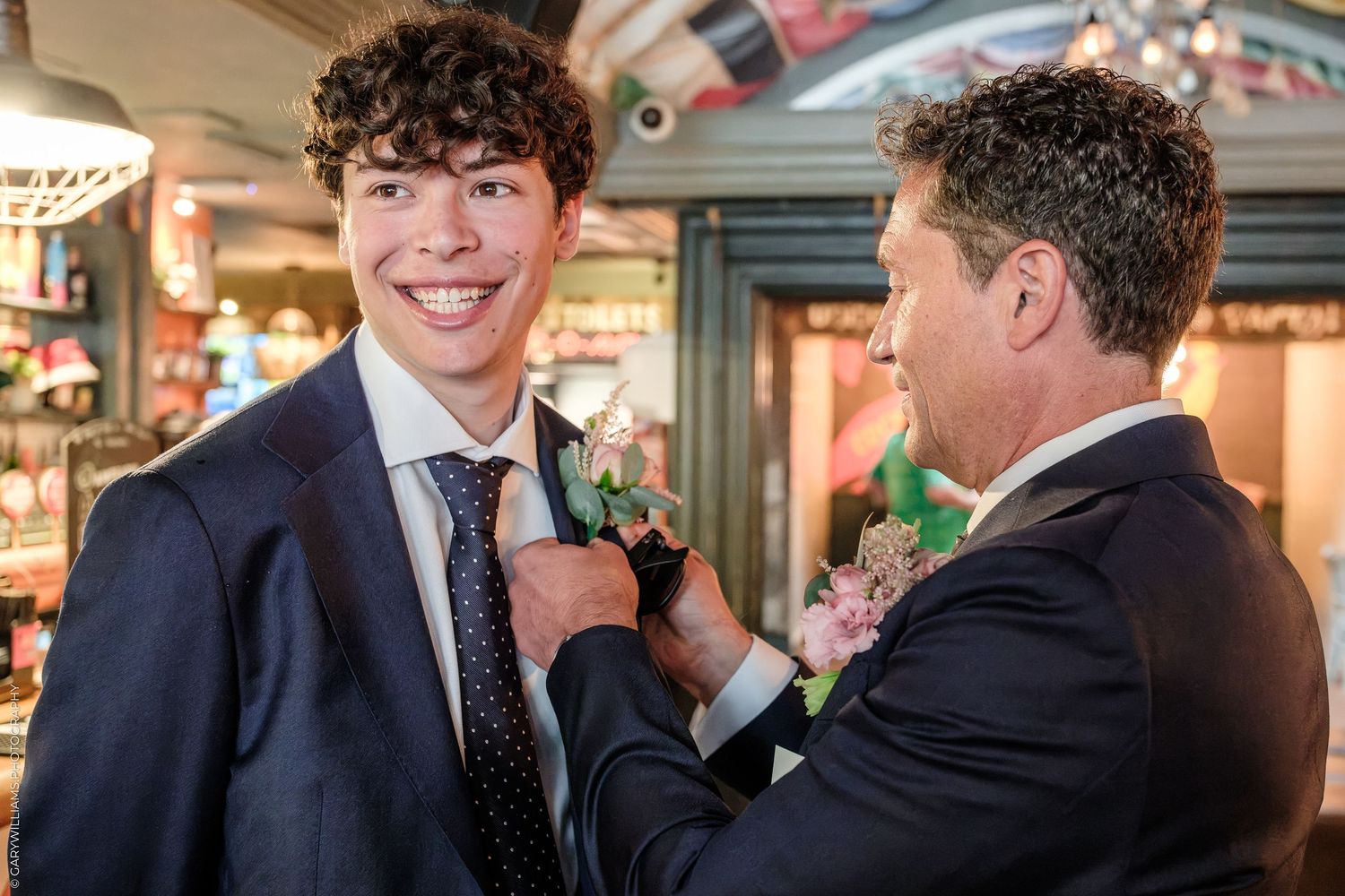 A man pins a boutonniere on a smiling young man in a suit and tie at a formal event.