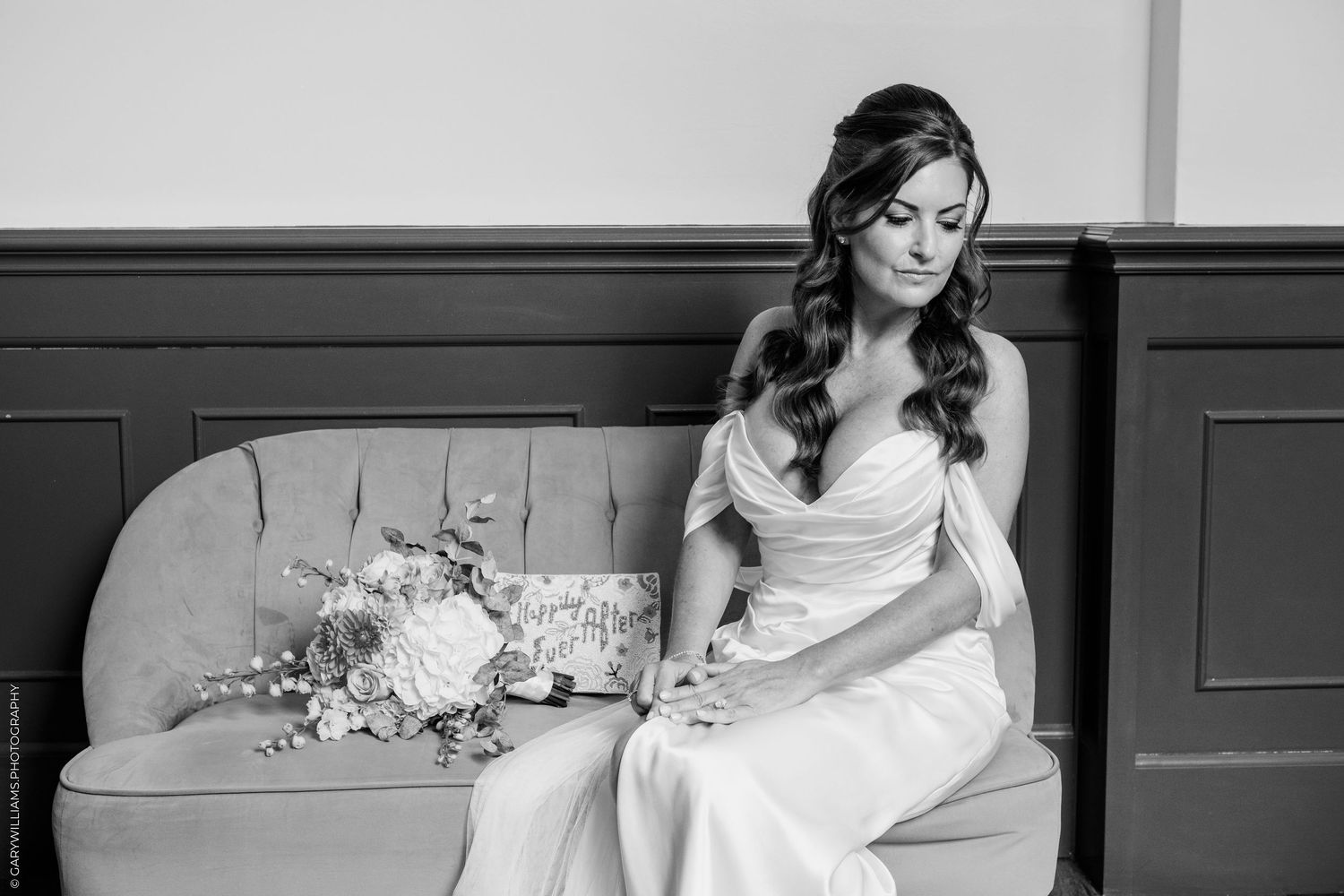 Bride in white gown sits on vintage sofa with floral bouquet, gazing down in elegant black and white wedding photo.