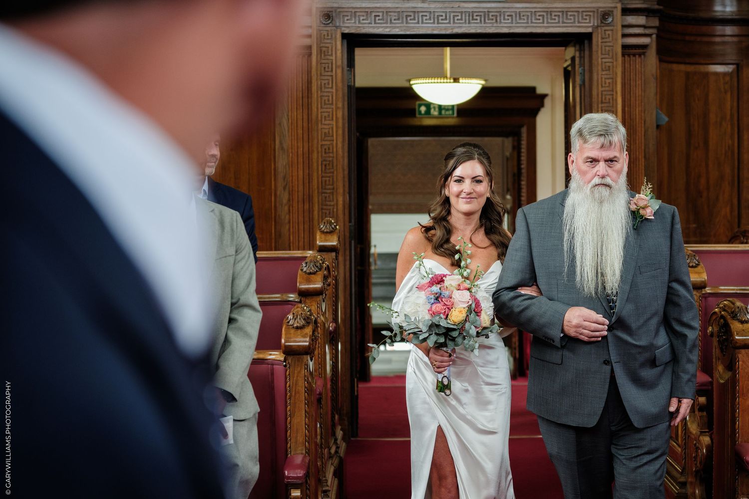 Bride walks down the aisle with bearded father at elegant wood-paneled wedding venue.
