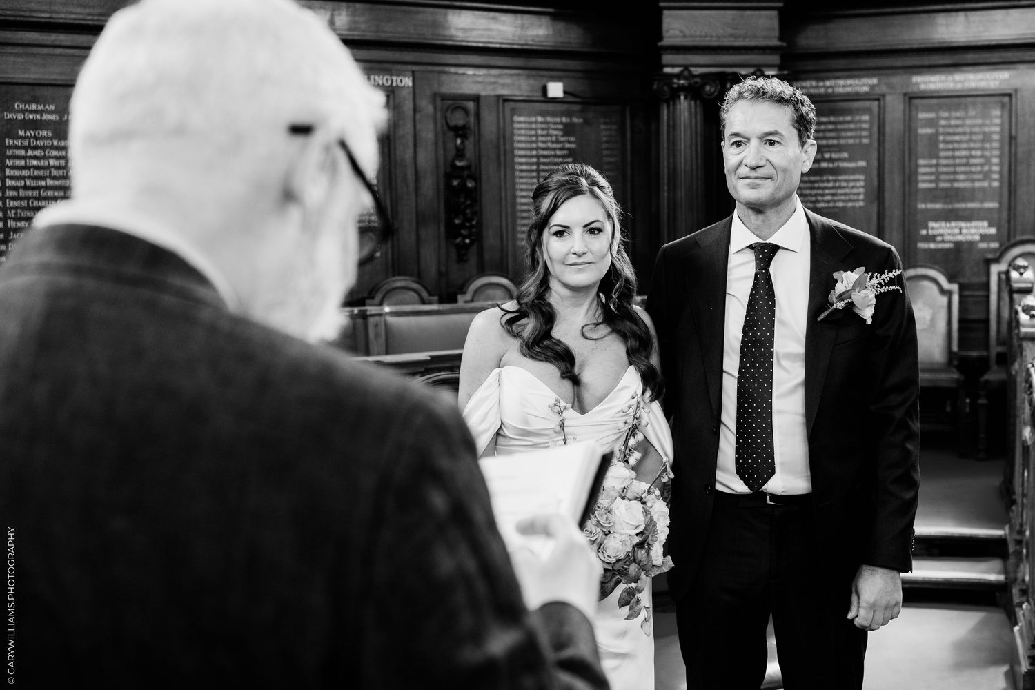 Black and white photo of a bride and groom standing before an officiant during an elegant indoor wedding ceremony.