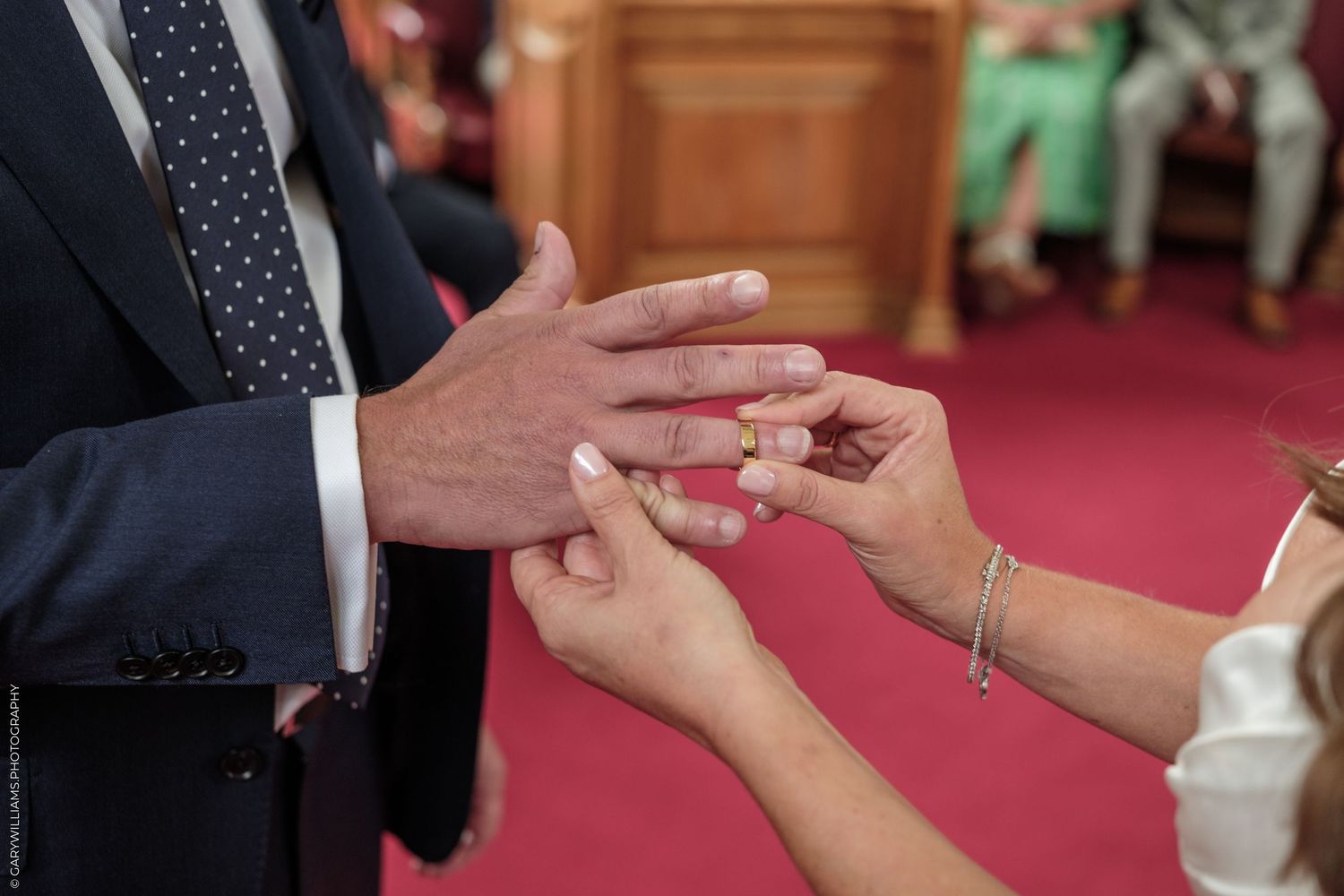 Bride placing a wedding ring on groom's finger during a ceremony, close-up of hands with bracelet.