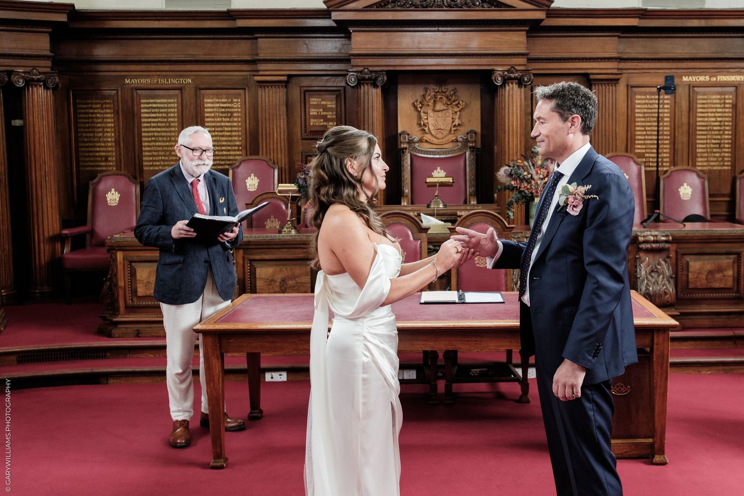 Couple exchanging vows at an elegant wood-paneled ceremony hall with an officiant reading from a book.
