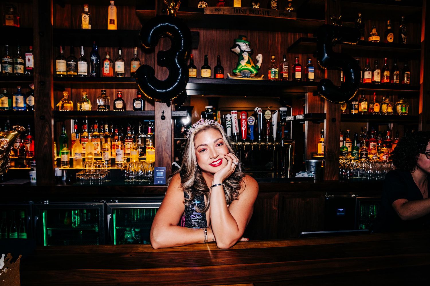 Smiling blonde woman leaning on a bar counter surrounded by shelves of liquor bottles in a dimly lit bar.