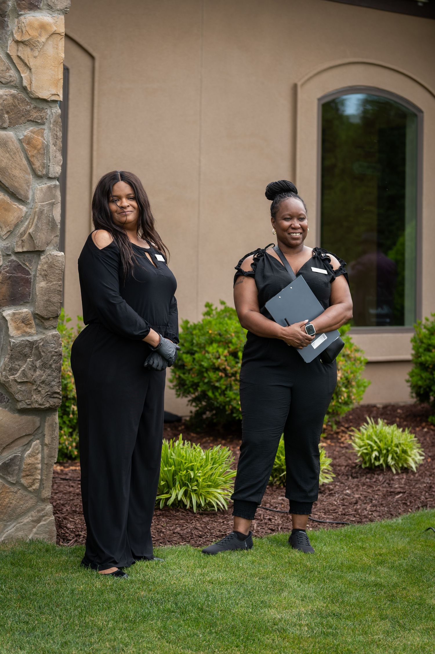 Two professional women in black outfits standing outside a building with stone accents and green landscaping.