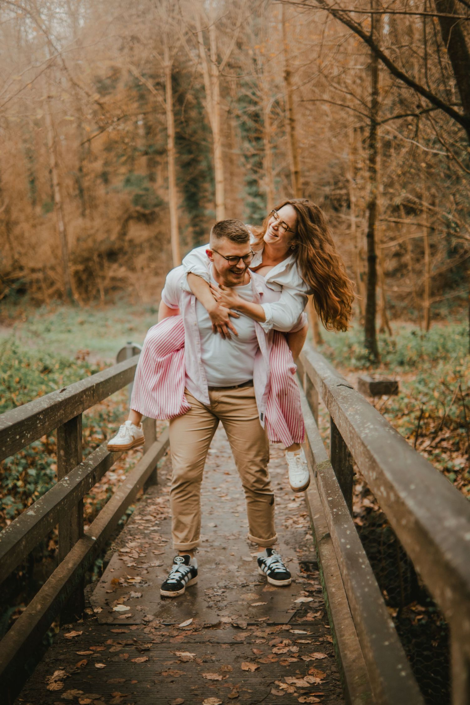 Happy couple sharing a piggyback ride on a wooden bridge surrounded by autumn forest foliage.