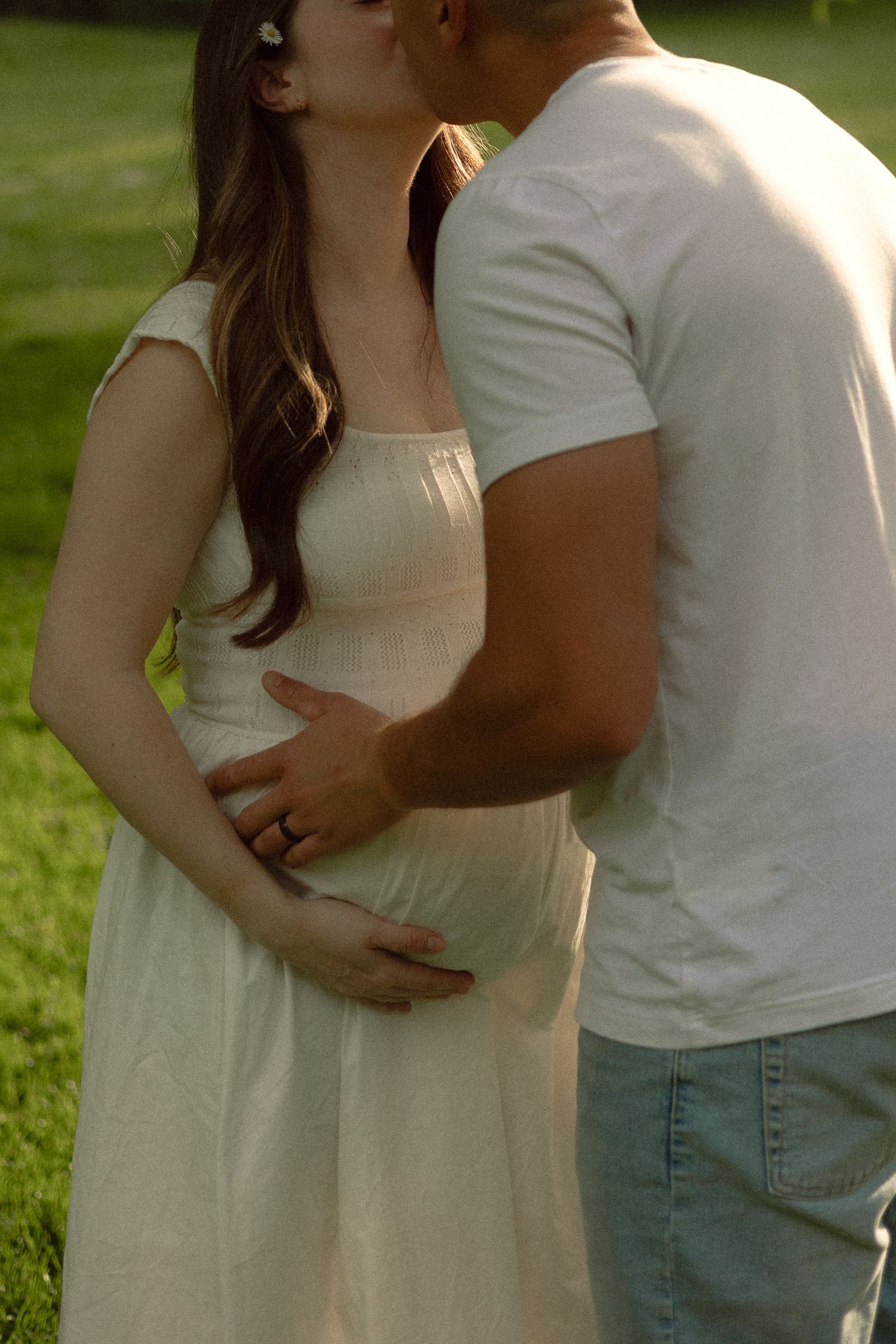 A couple shares a tender kiss outdoors, with the man gently cradling the pregnant woman's belly in a sunlit park.
