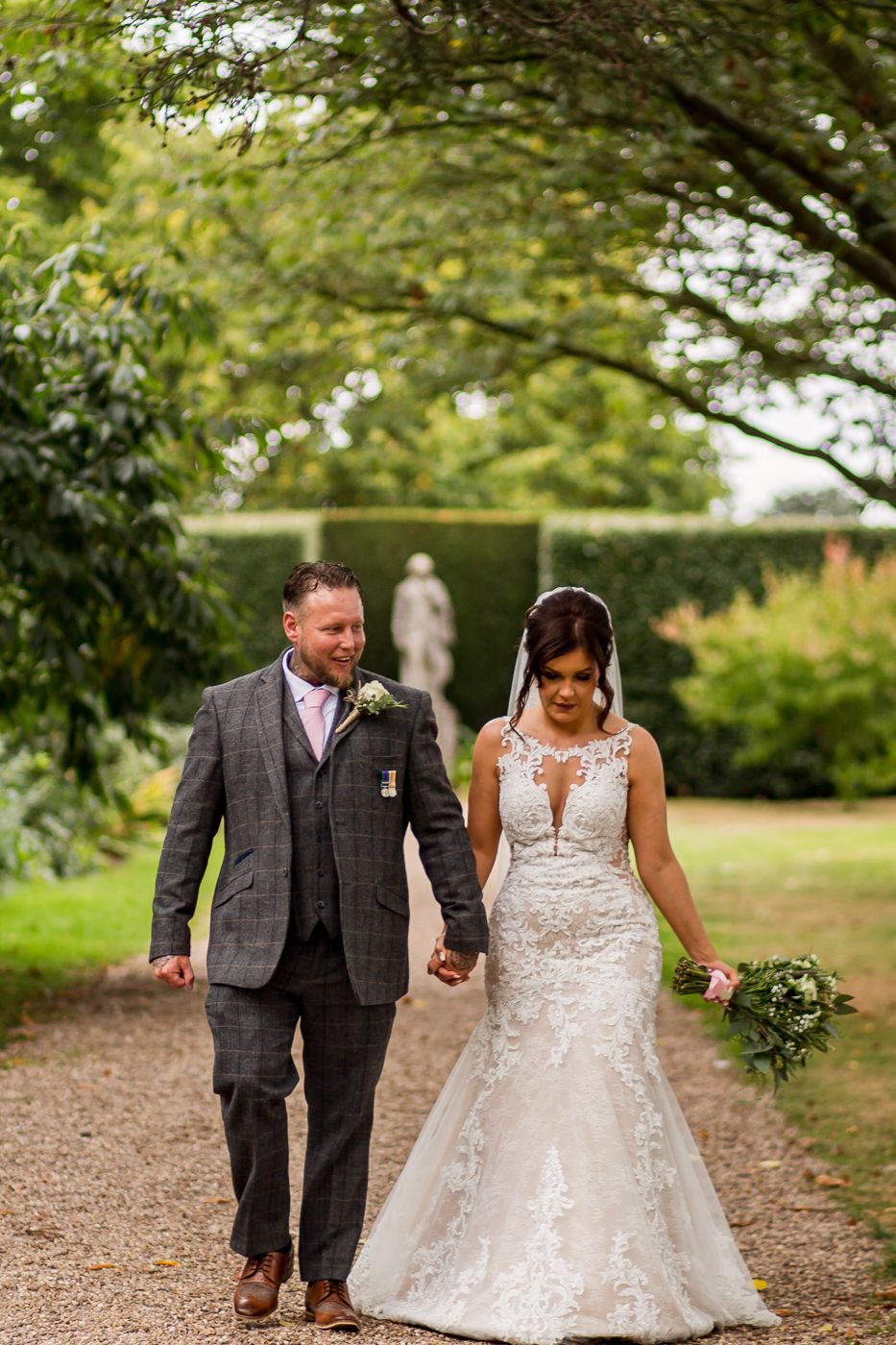 Newlyweds walk hand in hand along a tree-lined garden path.