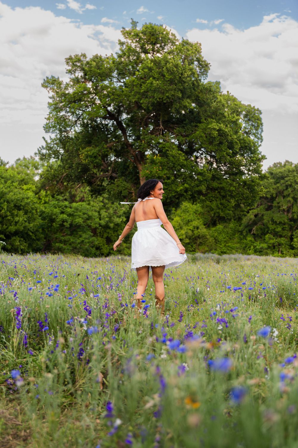 Woman in white dress walking through a field of blue wildflowers with large green trees and cloudy sky.