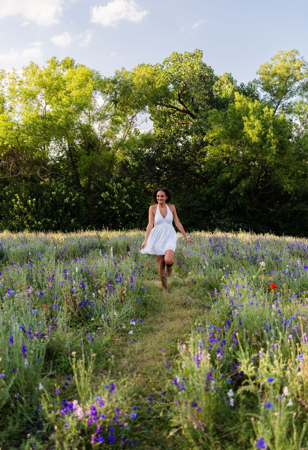Woman in white dress running through a vibrant wildflower field with purple blooms and lush green trees.