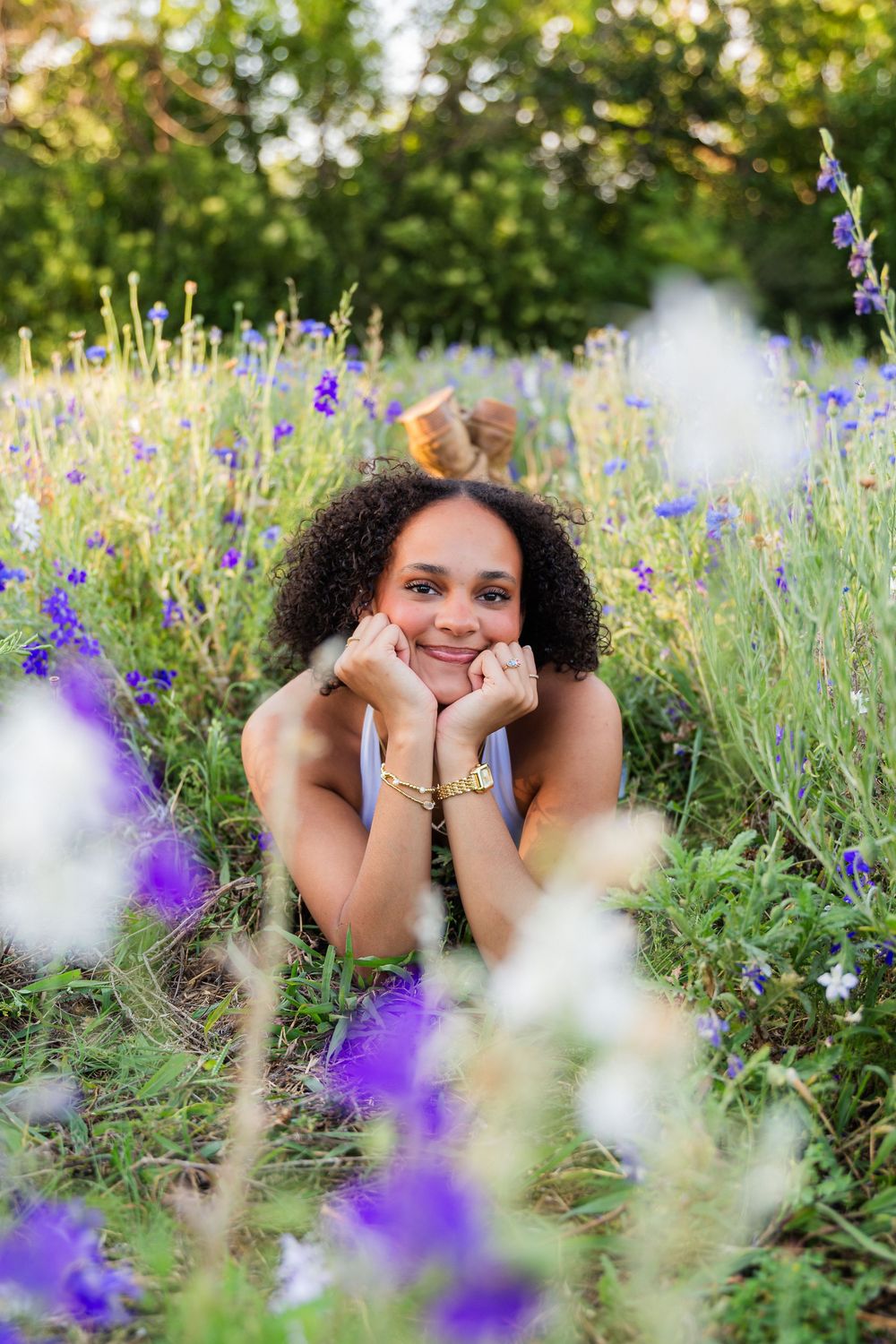 Young woman with curly hair lying in a wildflower field, smiling with hands on cheeks, surrounded by purple and white blooms.