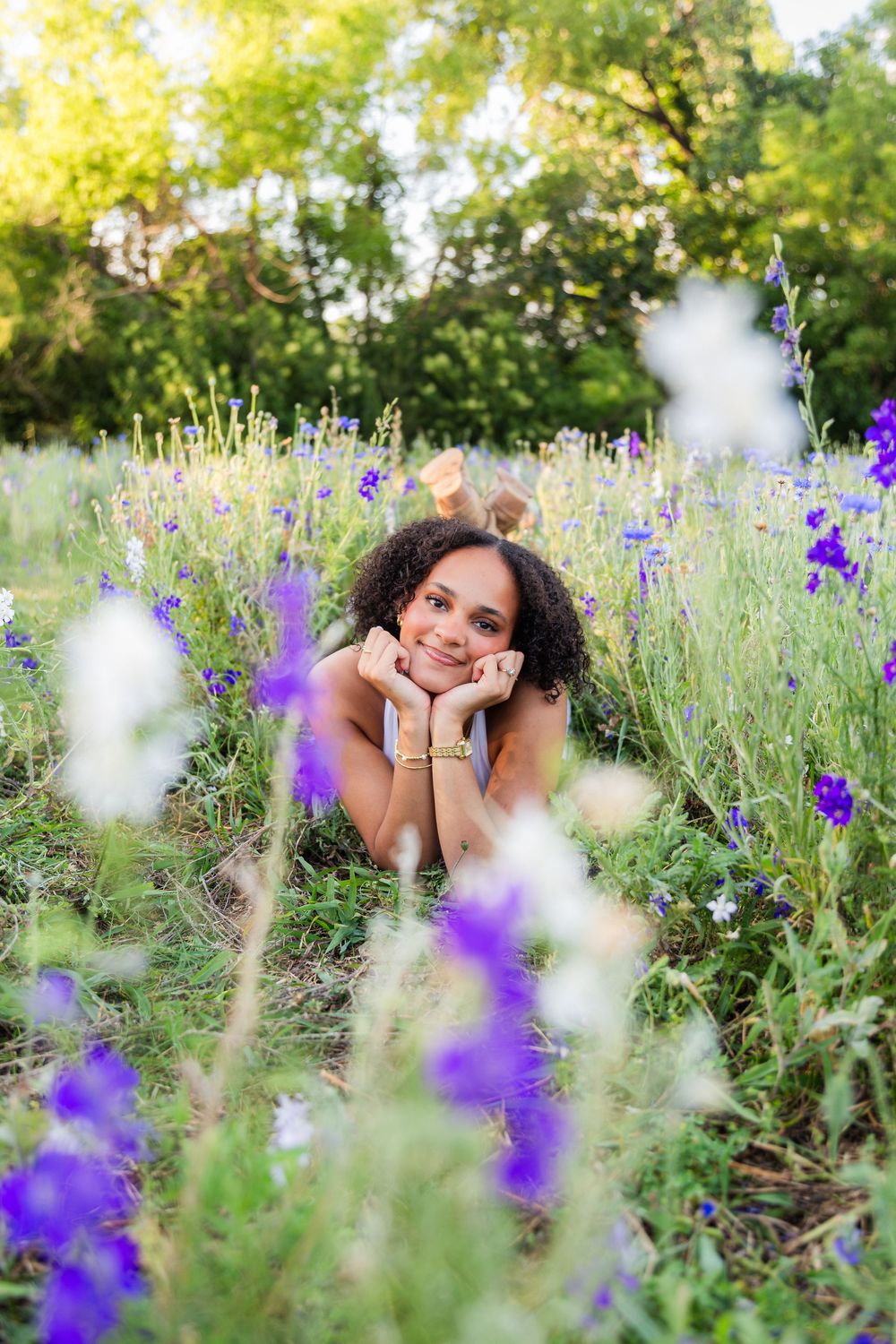 Young woman with curly hair lying in a field of purple and white wildflowers, smiling dreamily on a sunny day.
