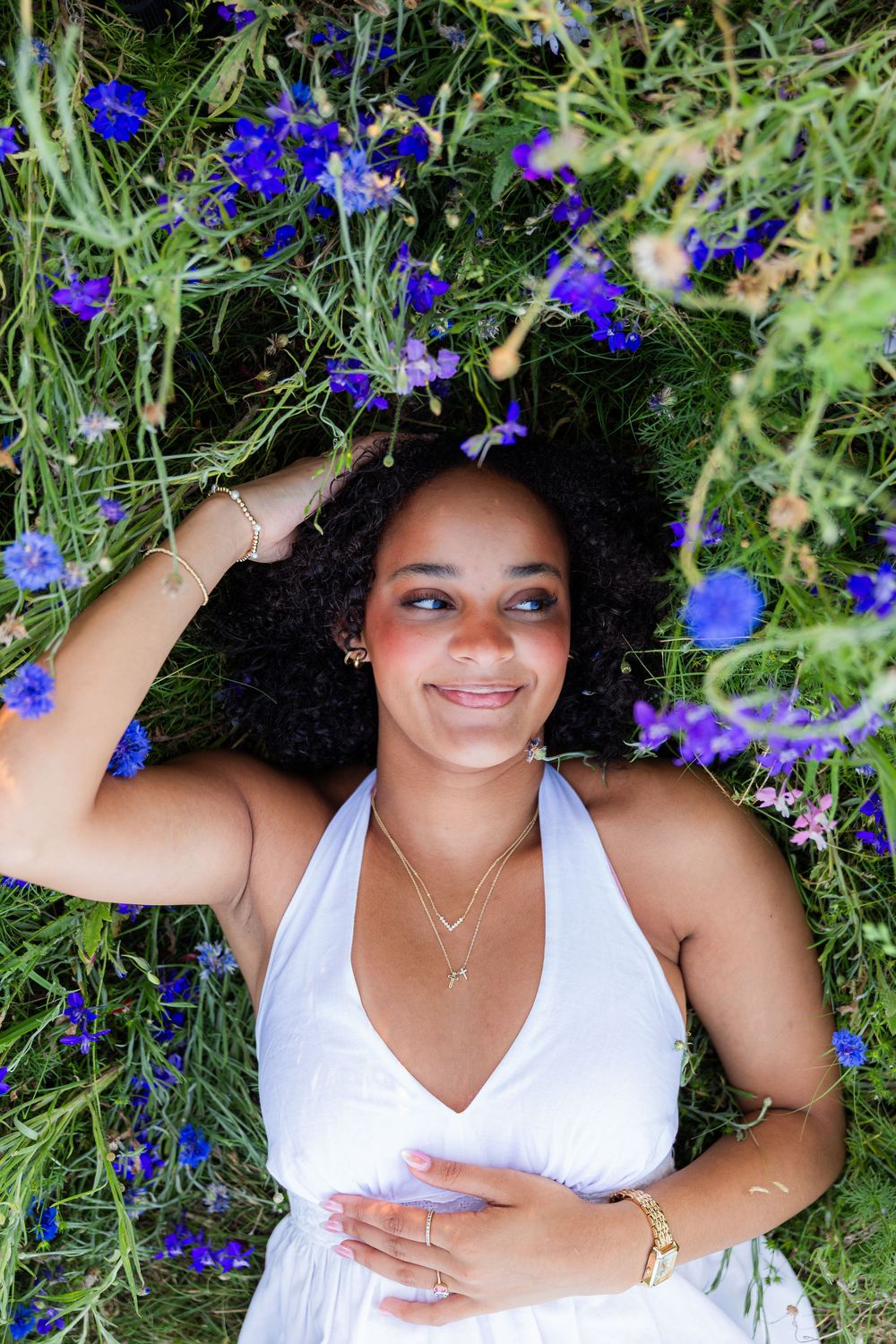 A smiling young woman in a white dress lies in a field of vibrant blue wildflowers, hand in her curly hair.