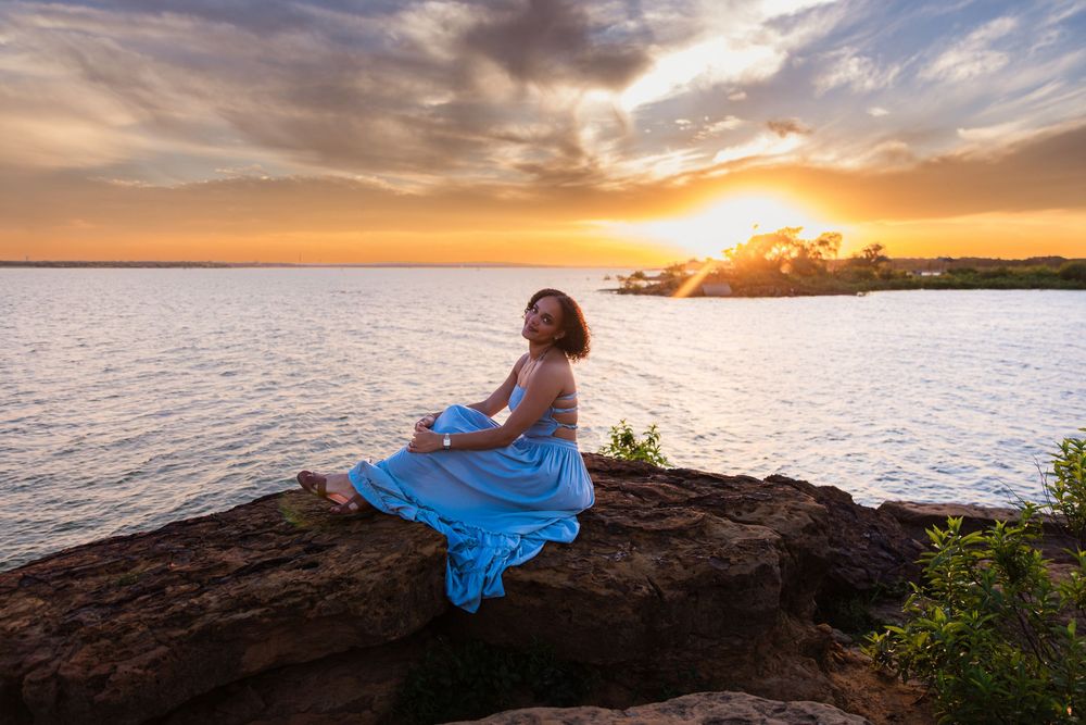 Woman in blue dress sitting on rocky cliff at golden sunset over calm ocean waters.