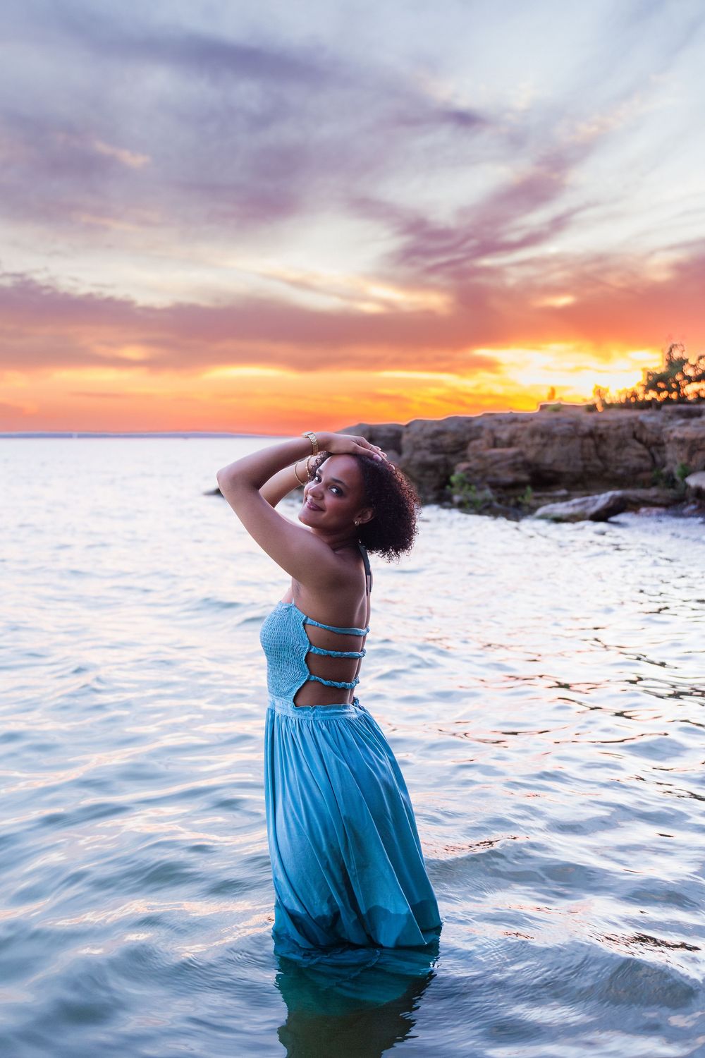 Woman in teal dress standing in ocean water at sunset with vibrant purple and orange sky backdrop.