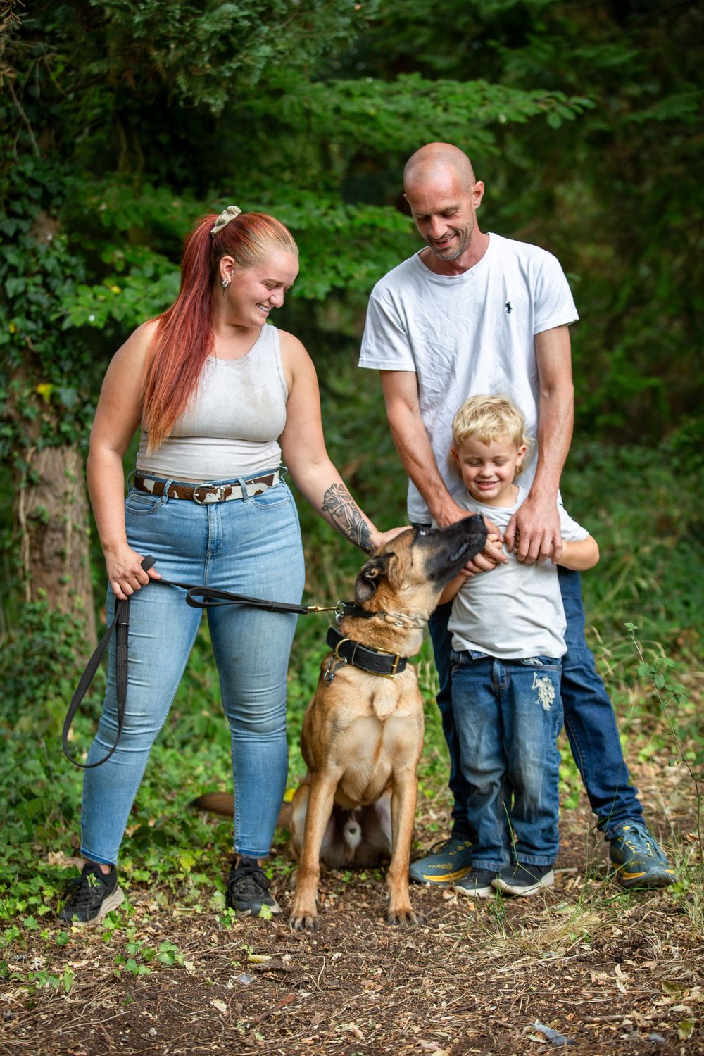 Family with red-haired woman, bald man, and blonde boy petting a German Shepherd dog outdoors in a forest setting.