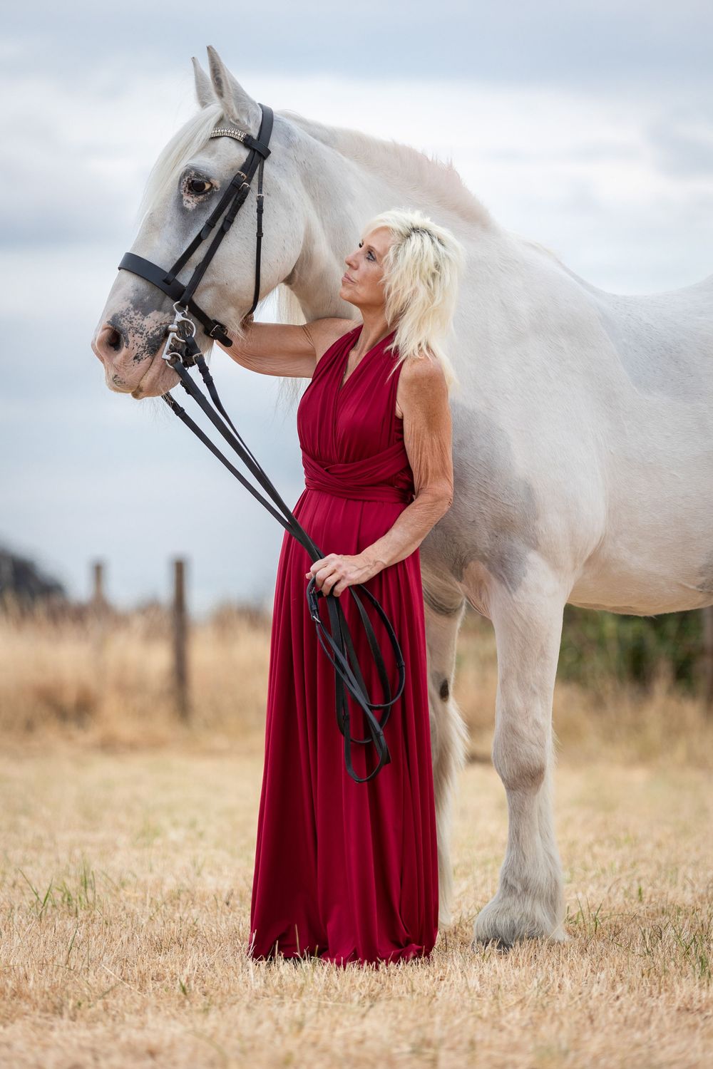 A blonde woman in a red gown stands beside a white horse in a golden field, gently holding its bridle.