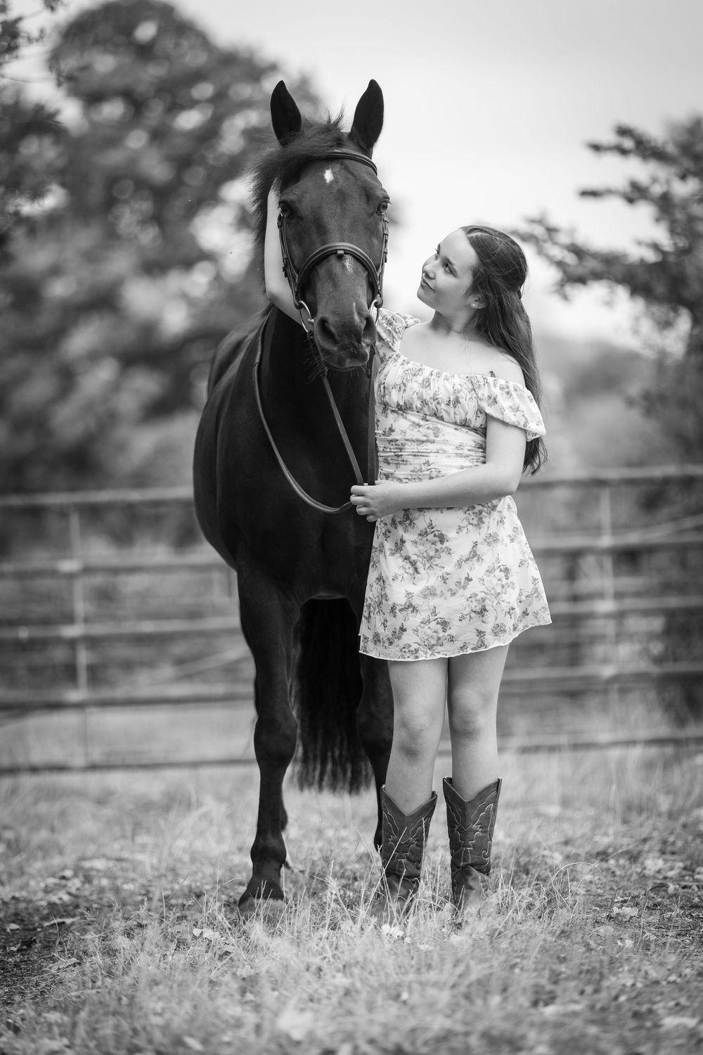 Black and white photo of a young woman in a floral dress and cowboy boots standing beside a horse in a fenced field.