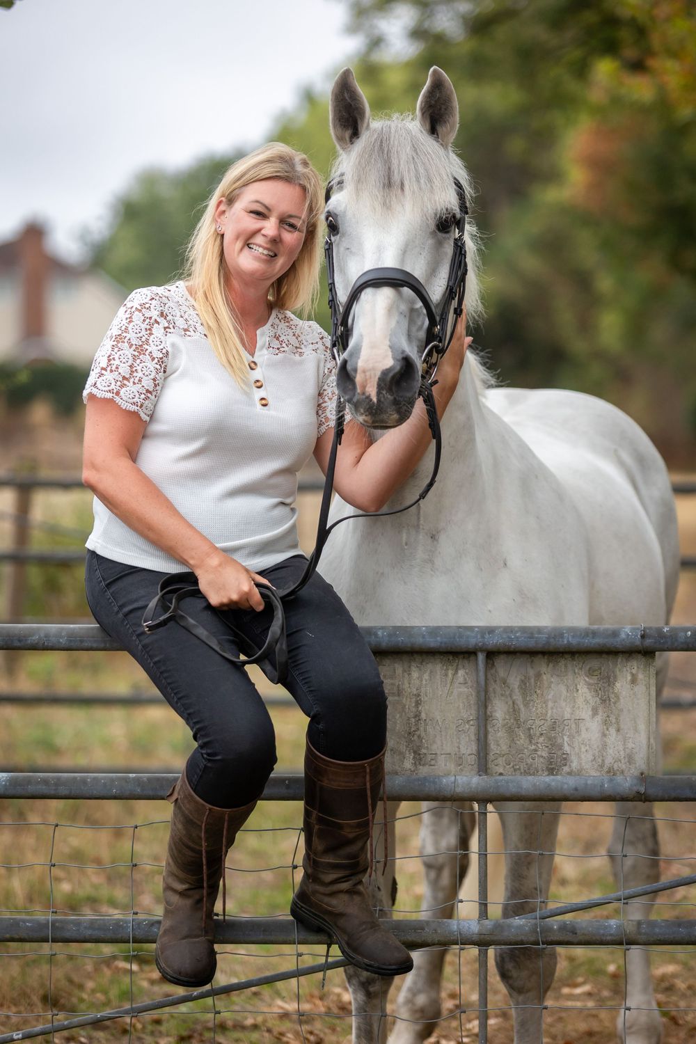 Smiling blonde woman in riding boots sits on a metal fence beside a white horse in an outdoor paddock.