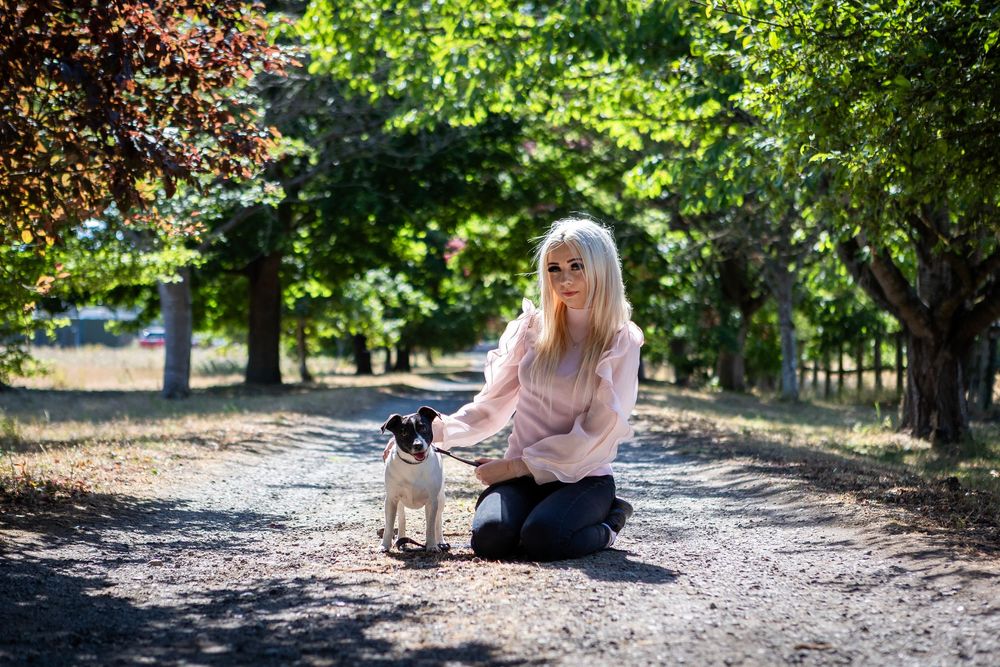 Blonde woman kneeling on a dirt path in a park, gently interacting with a small dog amid lush green trees.