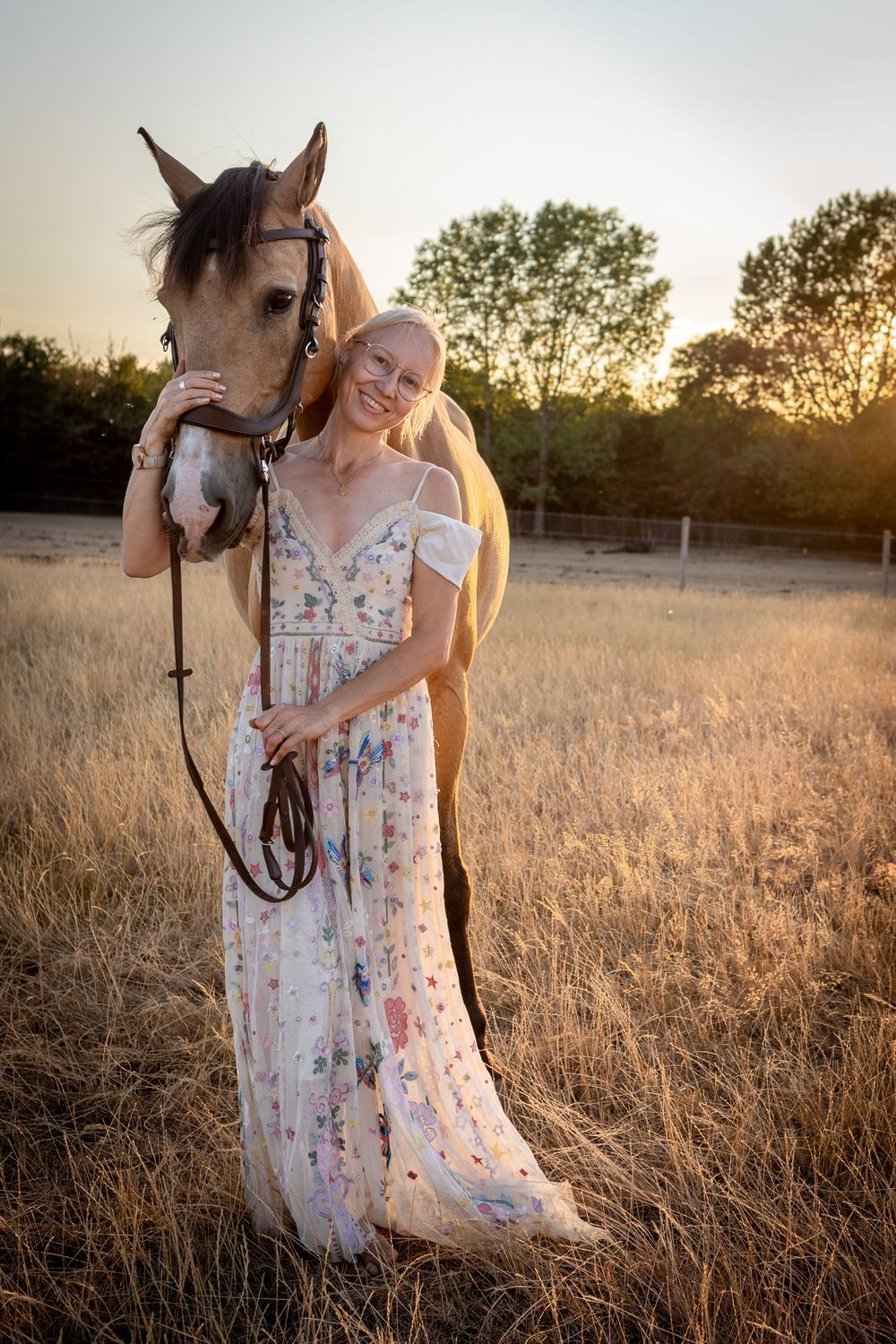 Woman in floral dress stands with brown horse in golden field at sunset.