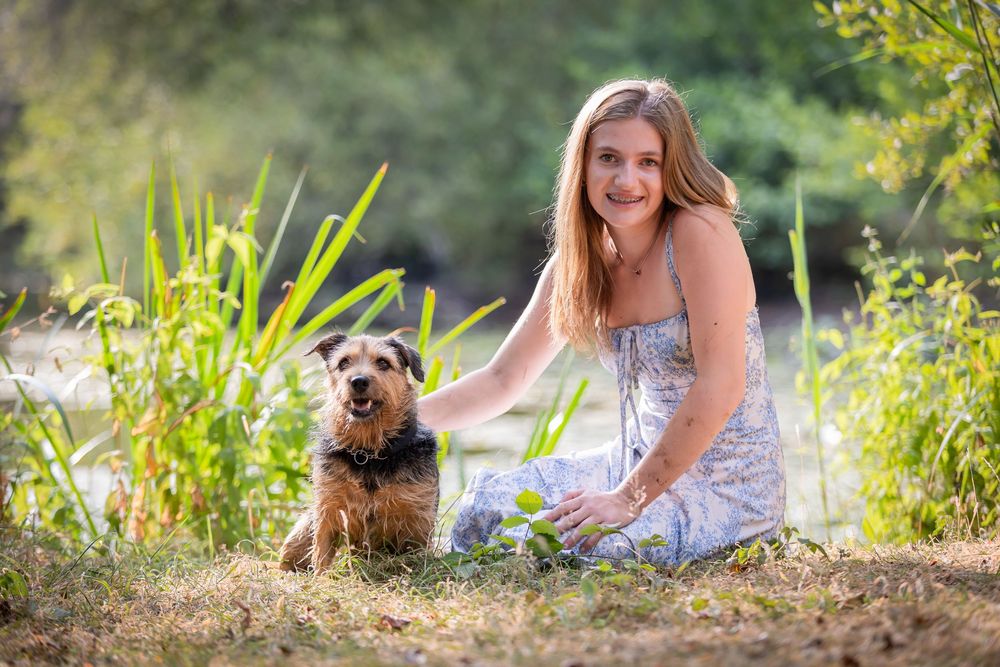 Young woman in floral dress smiling beside a terrier dog in a sunny outdoor setting near lush green plants.