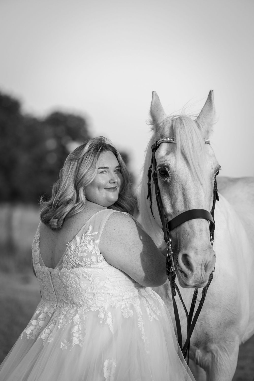 Black and white photo of a smiling bride in lace gown posing with a white horse in a field.