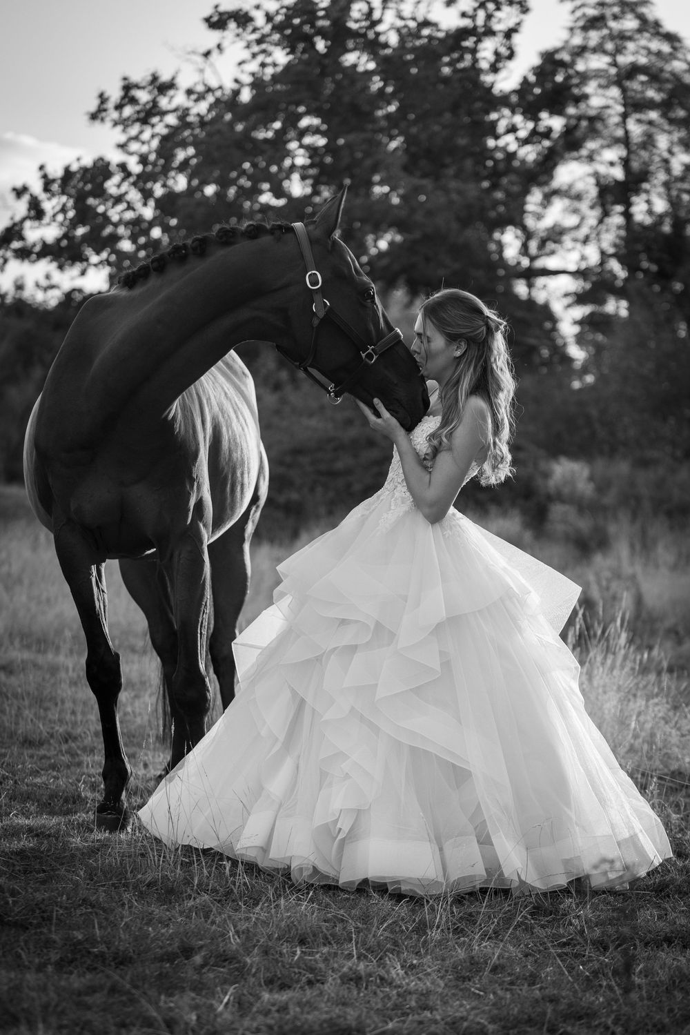 Black and white photo of a bride in a ruffled ball gown sharing a tender moment with a dark horse outdoors.
