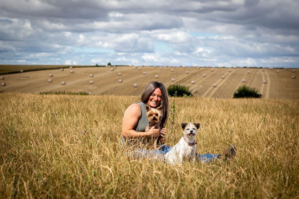 Woman sitting in golden wheat field with two small dogs, hay bales and cloudy sky in background.