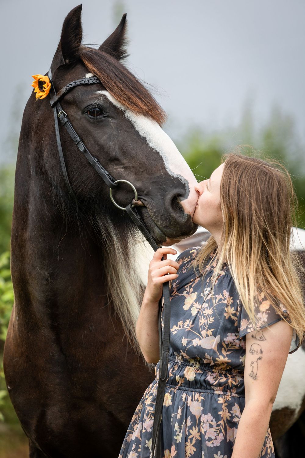 Young woman in floral dress kissing a dark brown horse with sunflower decoration on its bridle outdoors.