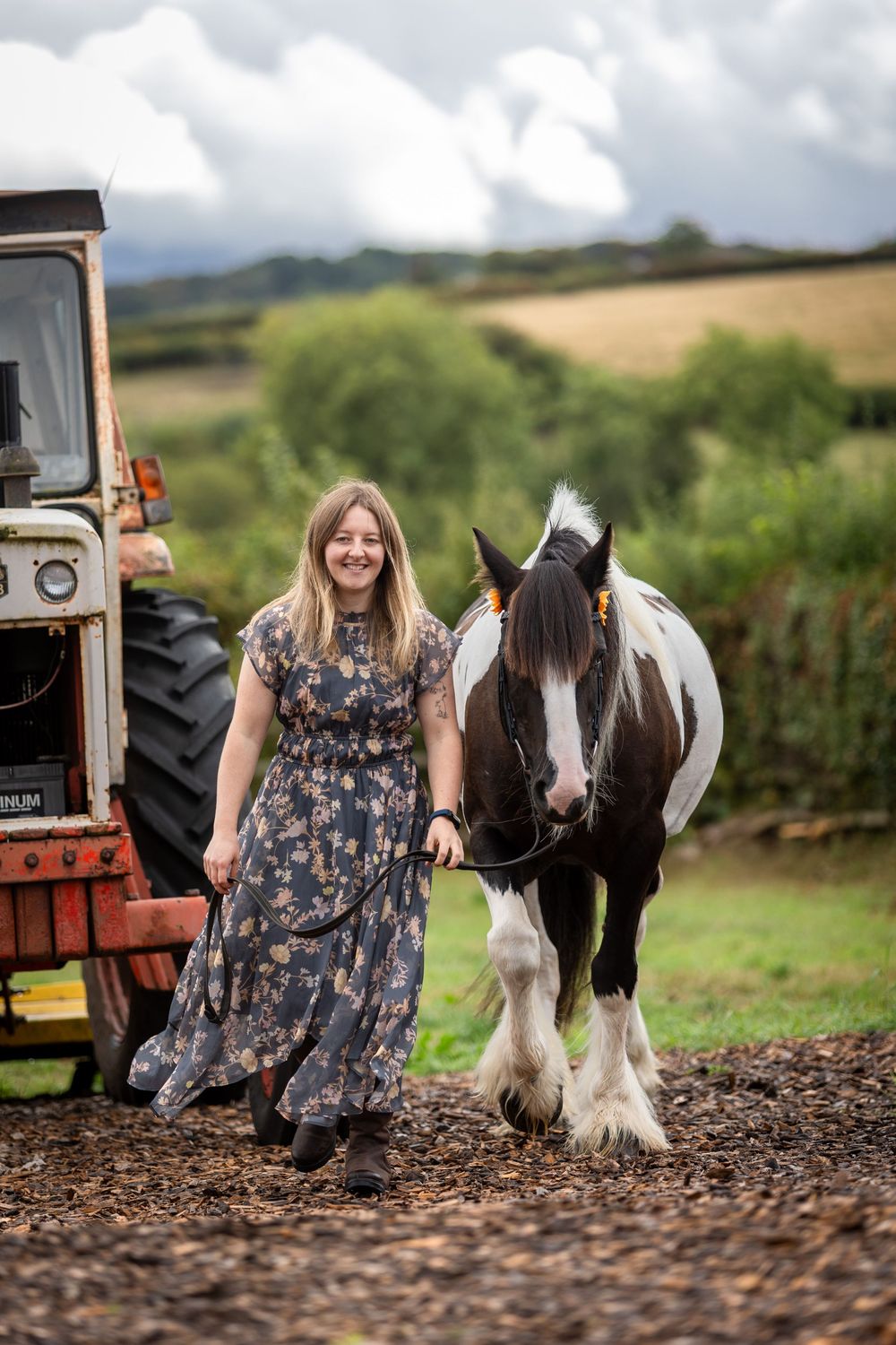 Woman in floral dress leading a piebald horse on a farm with a vintage tractor and green hills backdrop.