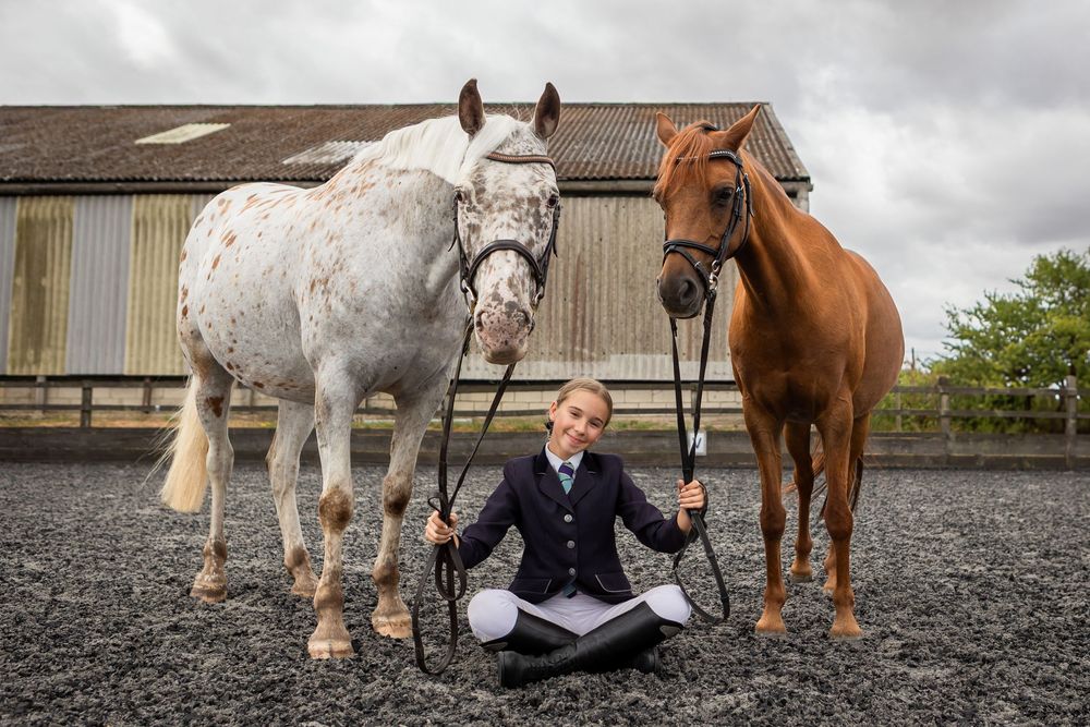 Young equestrian rider sits between two horses, a white Appaloosa and chestnut, holding reins in a stable yard.