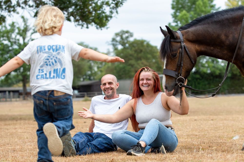 A family sits on grass with a dark horse nearby, as a child runs toward them outdoors.