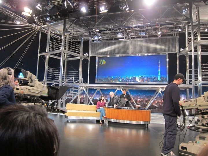 TV studio set with news desk, large LED screen displaying city skyline, camera equipment, and production crew on floor.