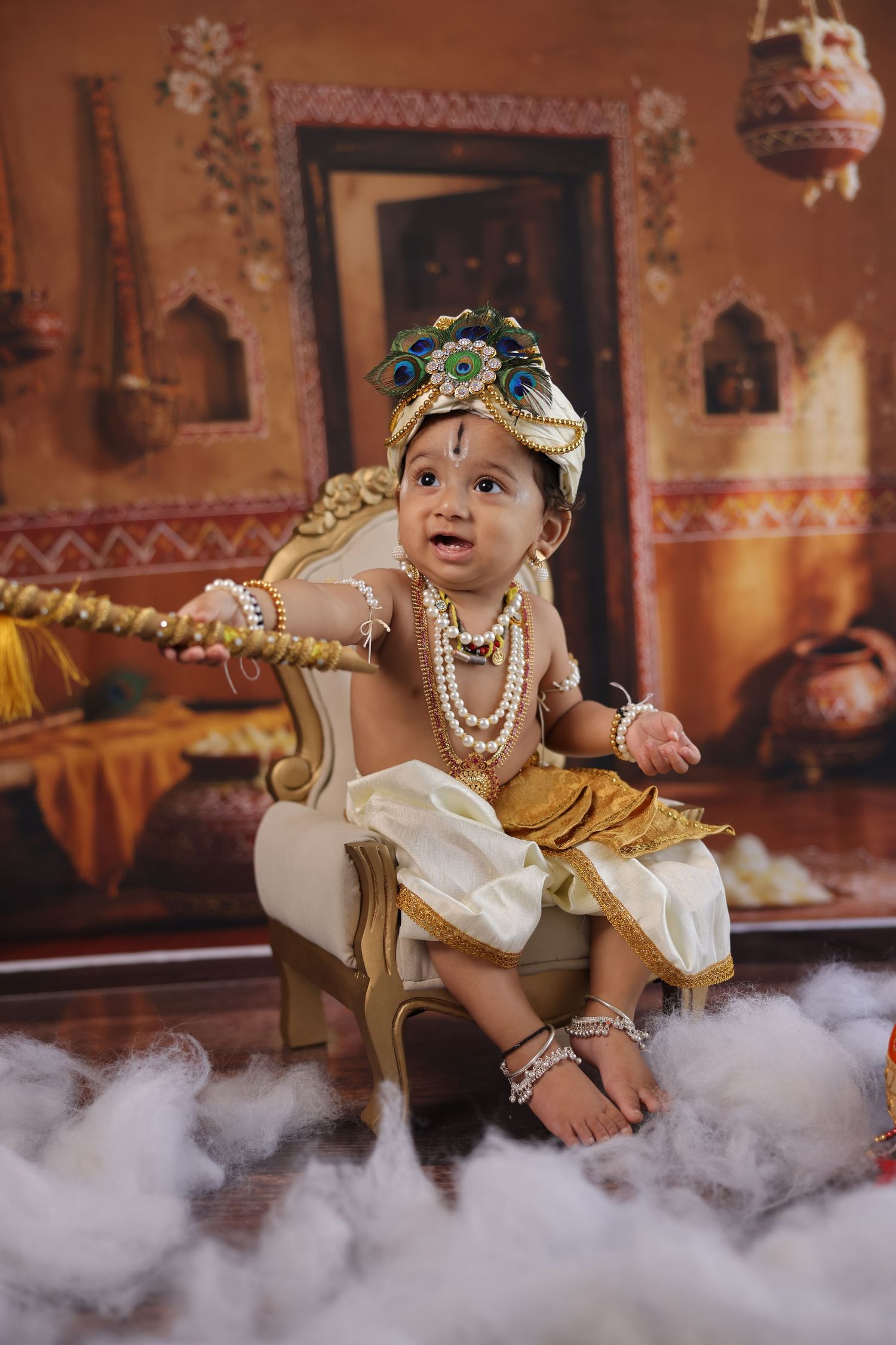 Baby dressed as Lord Krishna in gold outfit with peacock feather crown and pearl necklaces, seated on ornate chair.