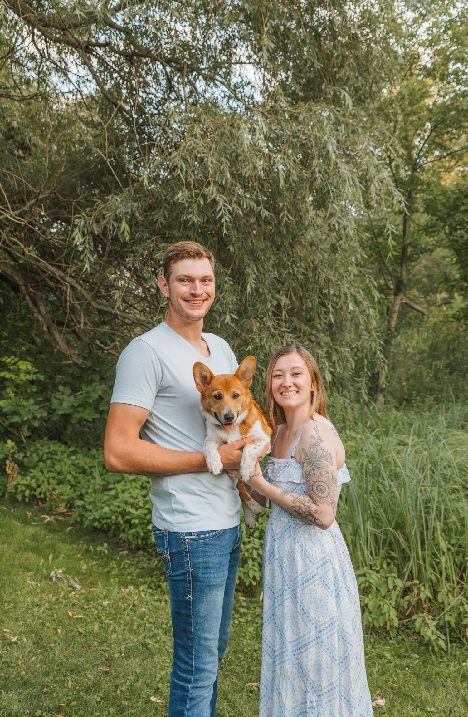 A smiling couple holds an adorable Corgi dog outdoors in a lush green garden setting.