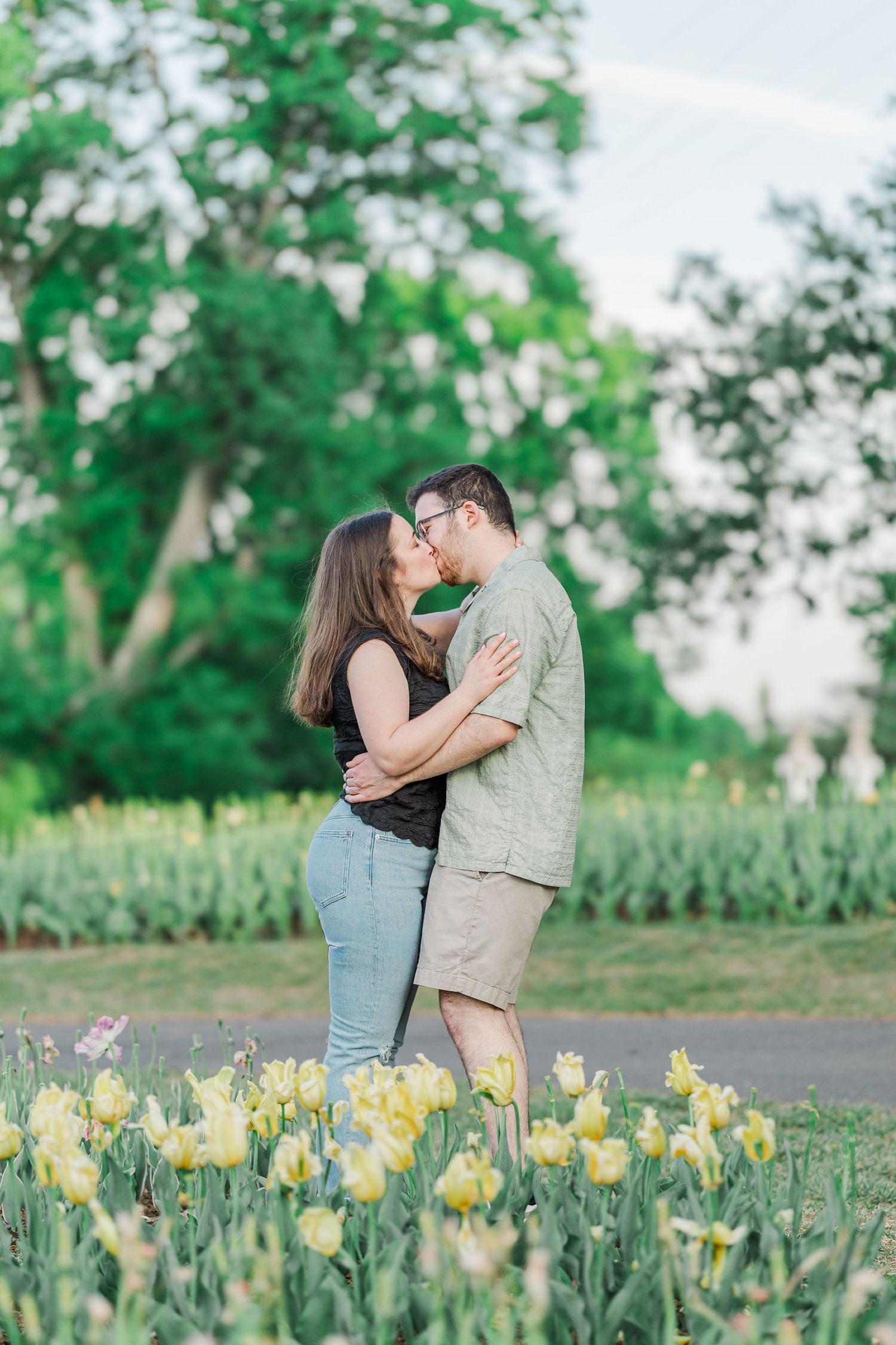 Couple kissing in a garden surrounded by yellow tulips and lush green trees during golden hour.